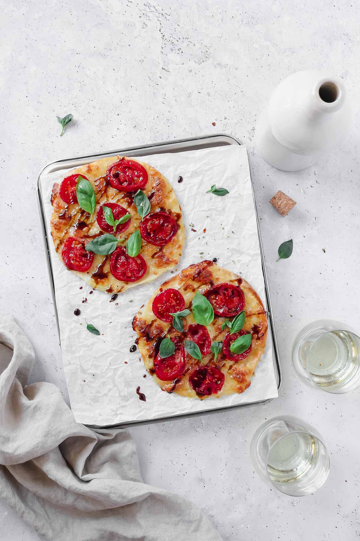 caprese tortilla pizza on baking sheet with parchment paper on grey background with wine glass and linen