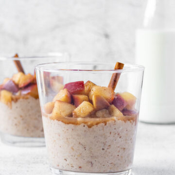 apple cinnamon oatmeal with cinnamon stick in tumbler glasses with milk standing behind on light grey background.
