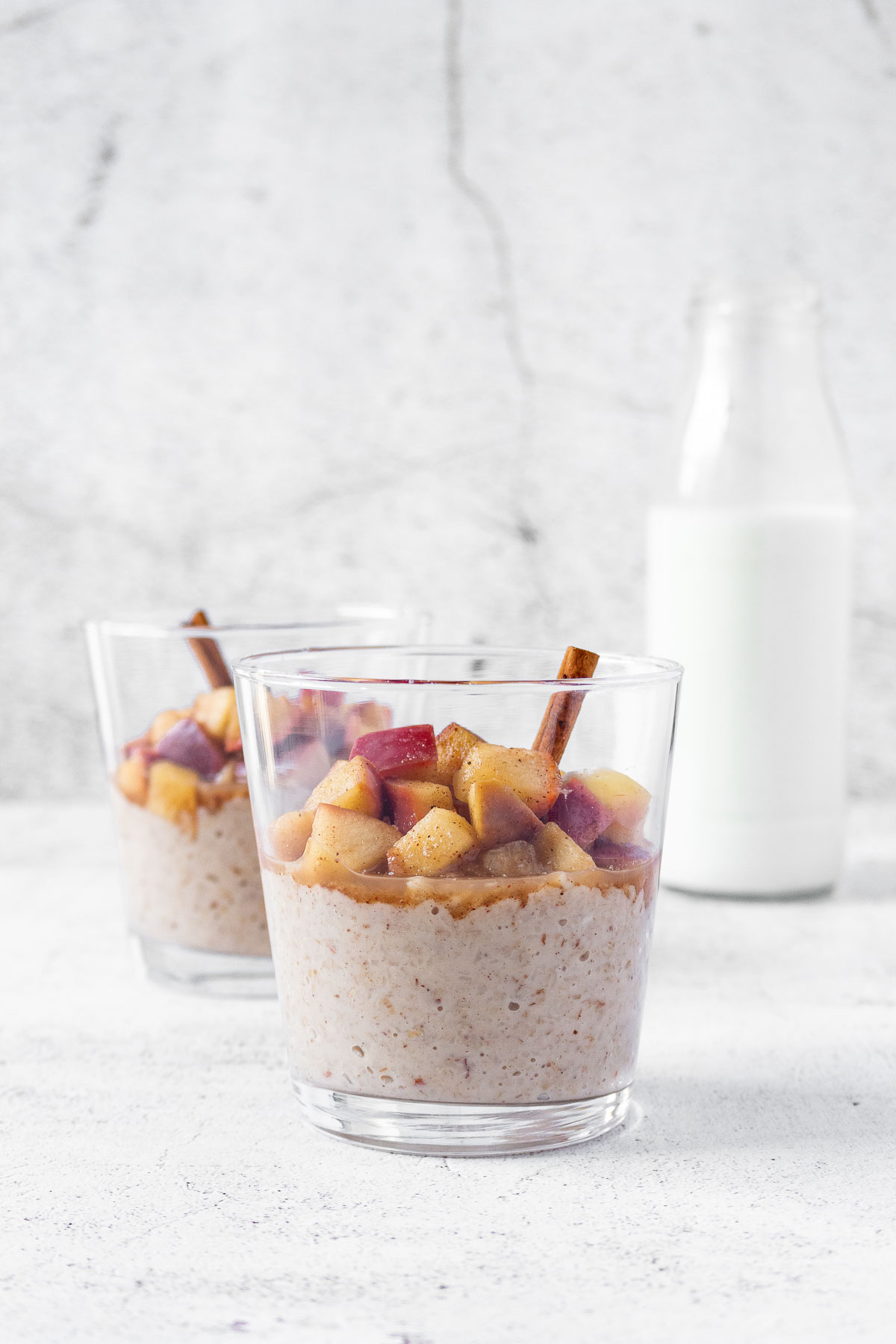 apple cinnamon oatmeal in tumbler glasses with milk standing behind on light grey background.