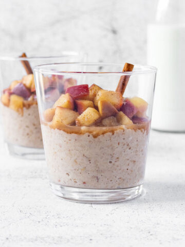 apple cinnamon oatmeal with cinnamon stick in tumbler glasses with milk standing behind on light grey background.