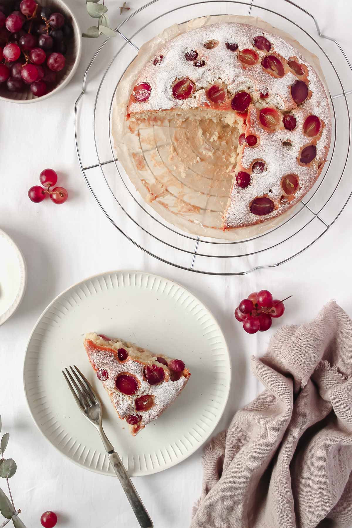 grape cake on cooling rack with grape decoration and white plate with piece of cake next to linen.