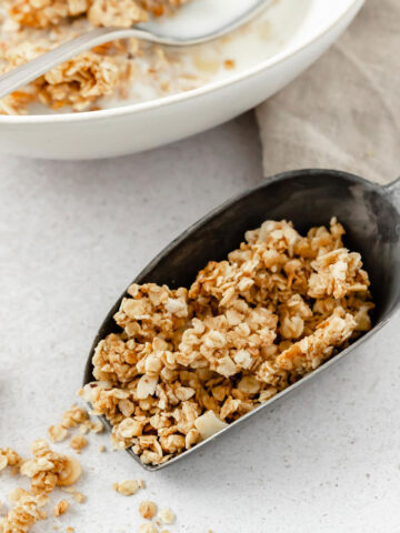 homemade granola in serving shovel with granola and milk bowl in the background.