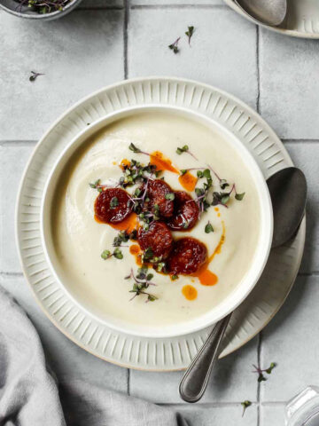 creamy cauliflower soup with crispy chorizo chips, herb and oil topping on grey tile background.