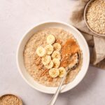 banana porridge with toppings on white bowl with vintage spoon and props on rosy background.