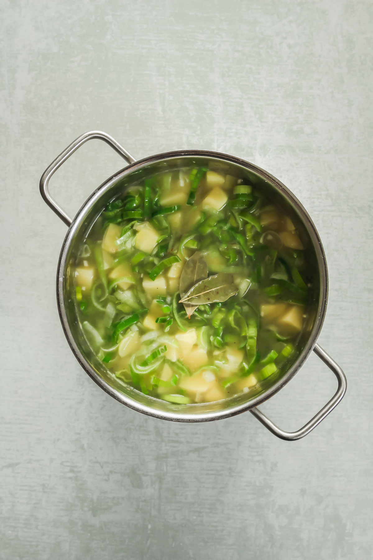 potatoes, leek, onion, garlic, bay leaves and vegetable stock in pot on green background.