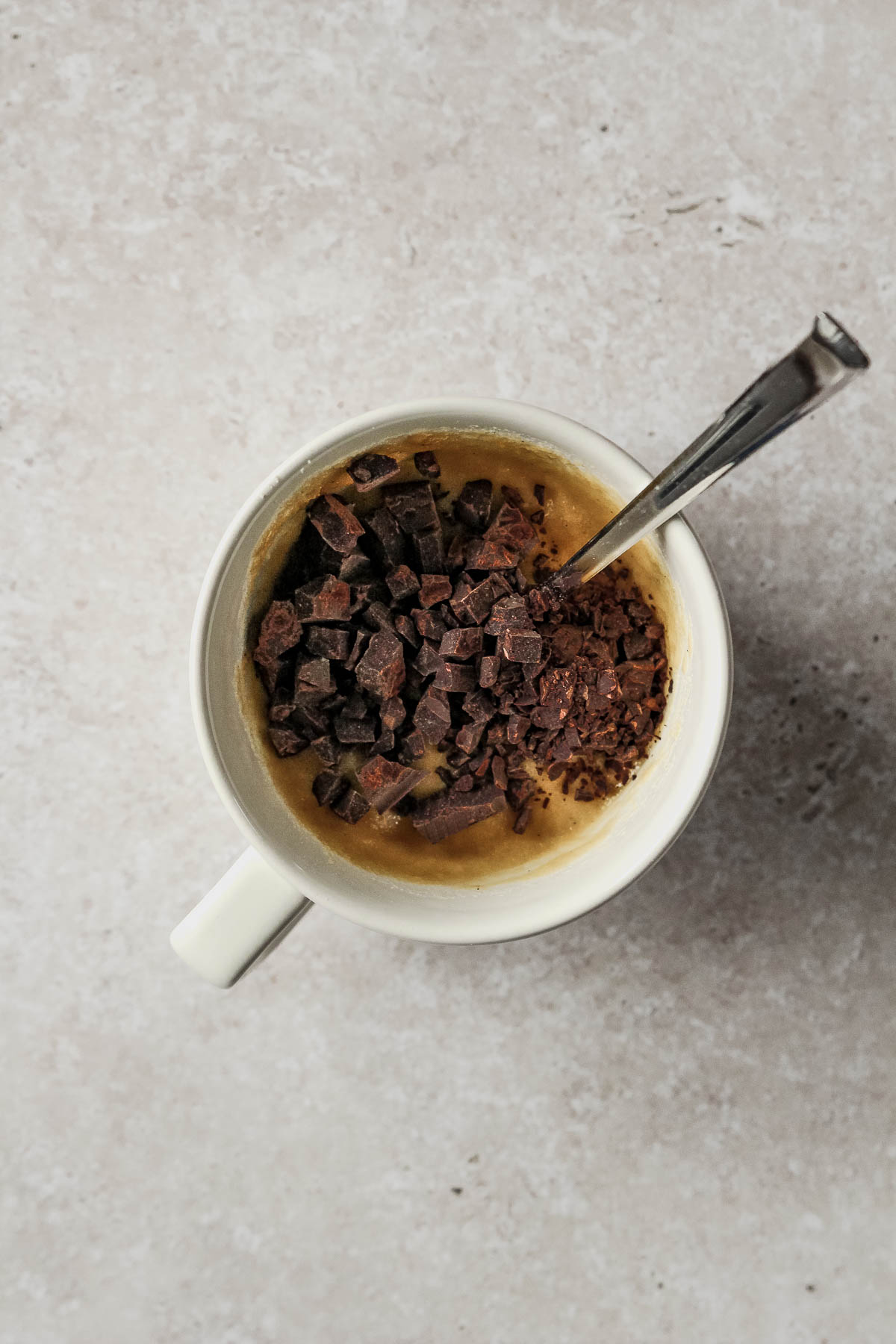 chopped dark chocolate about to be mixed in dough for chocolate almond flour mug cake in off-white mug on beige marble background.