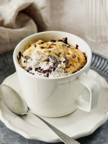 almond flour mug cake with chopped dark chocolate in off-white mug on white and vintage mental plat with vintage spoon.