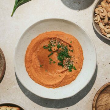 harissa hummus in beige bowl with crackers and nuts on beige marble background.