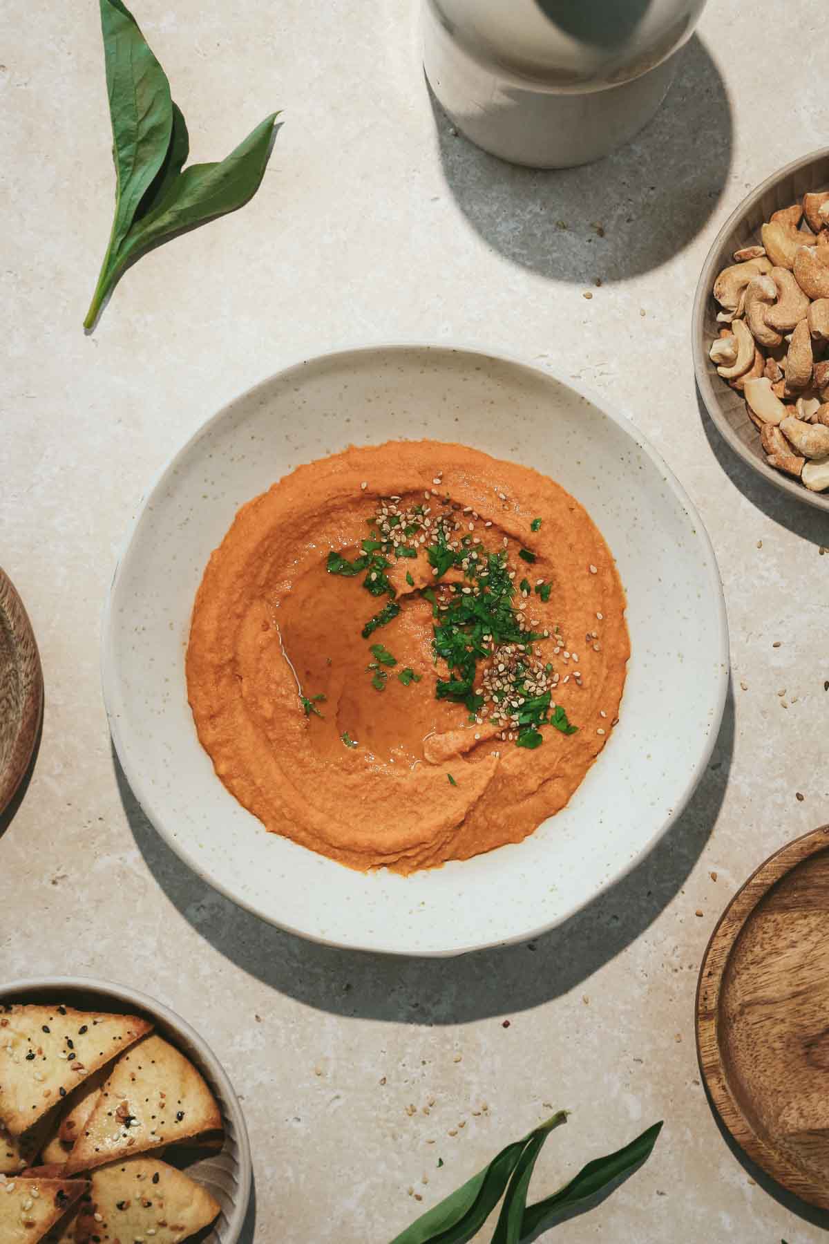 harissa hummus in beige bowl with crackers and nuts on beige marble background.