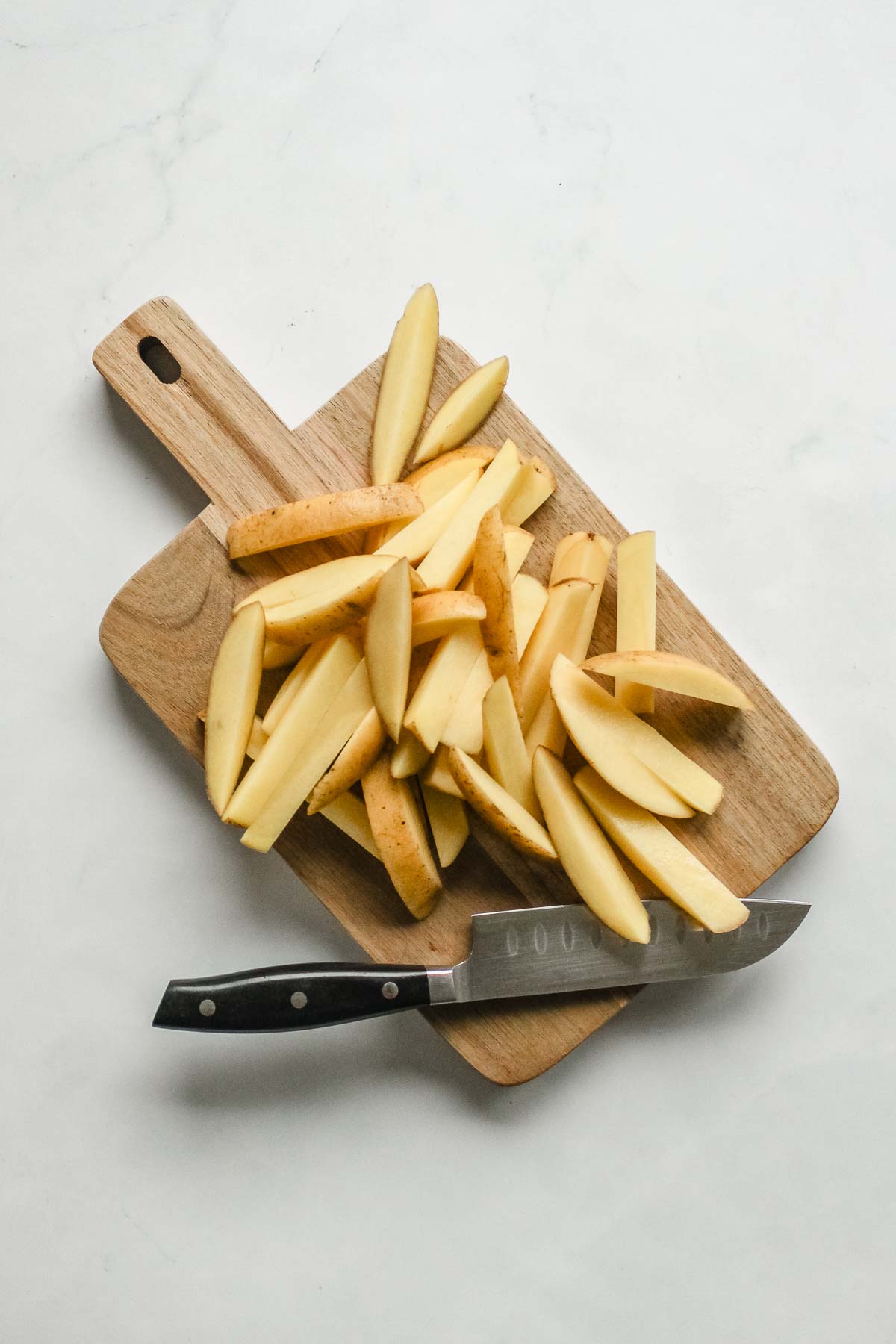 potatoes with skin cut into fries on wooden board with knife.