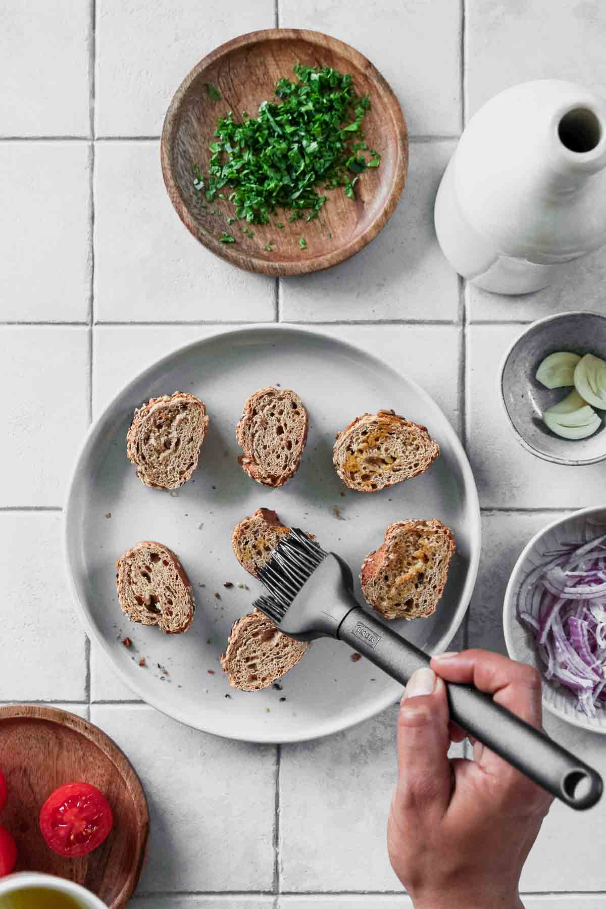 brushing olive oil on whole-wheat baguette slices with other ingredients in background.