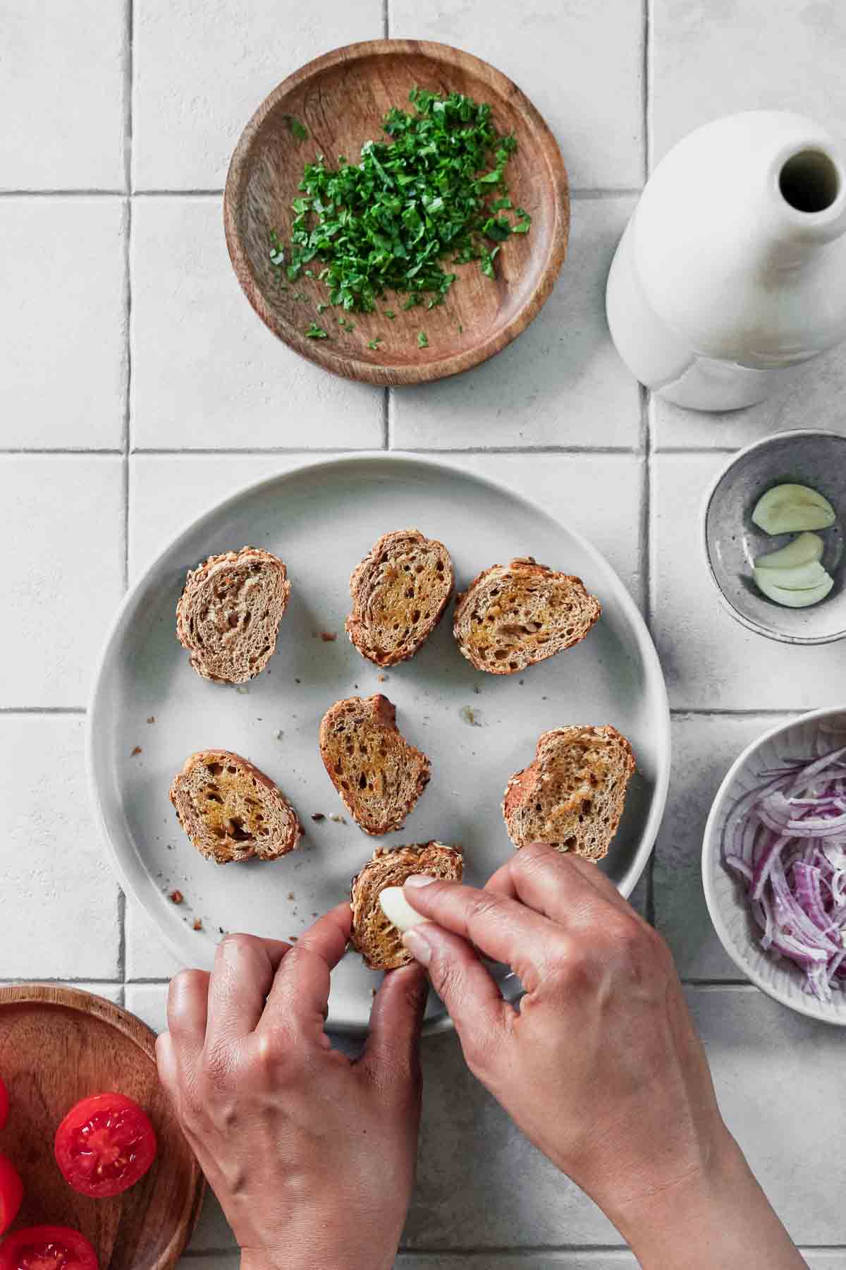 rubbing garlic on whole-wheat baguette slices with other ingredients in background.