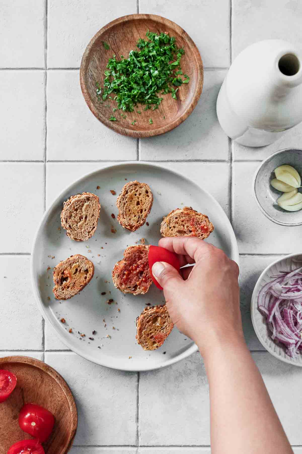 rubbing halved tomato on whole-wheat baguette slices with other ingredients in background.