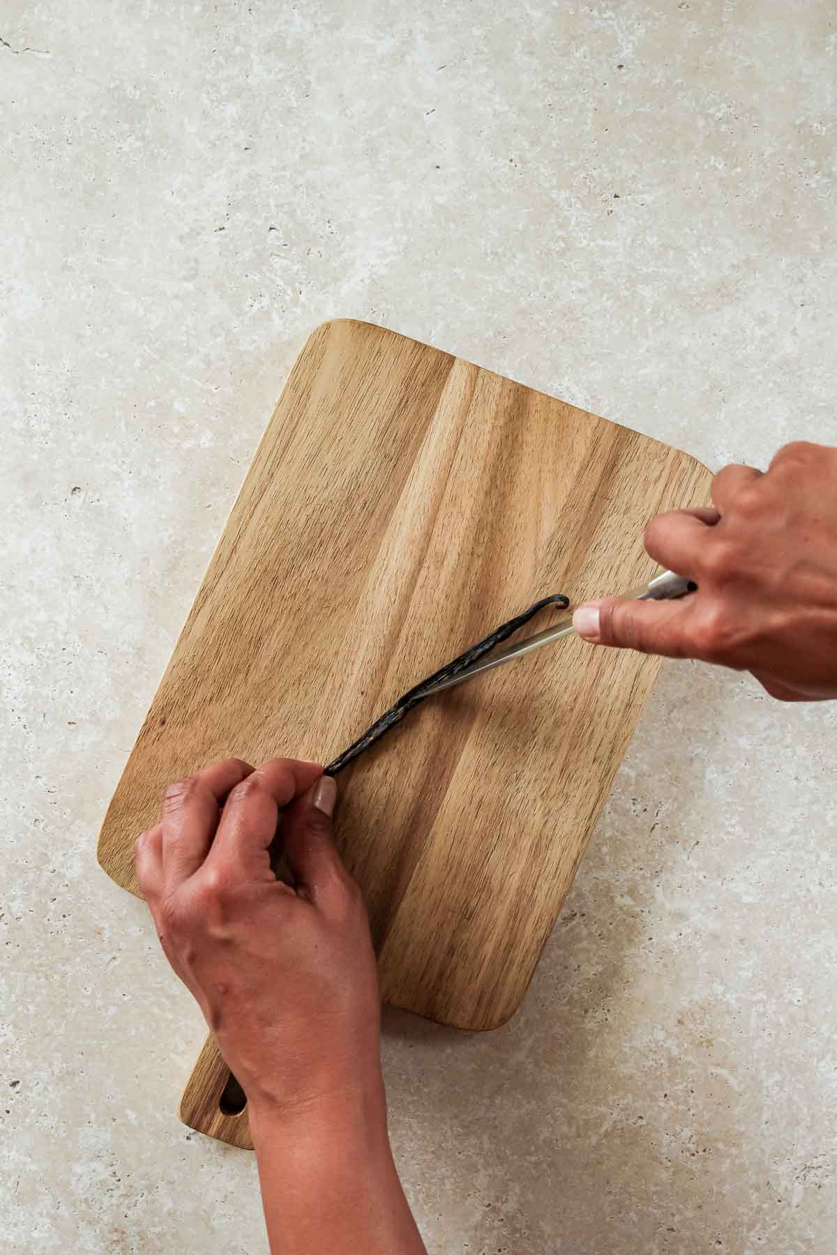 slicing vanilla bean open with a knife on wooden cutting board.