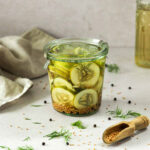 sweet and sour pickled cucumber sin glass jar with herbs and spices scattered around.