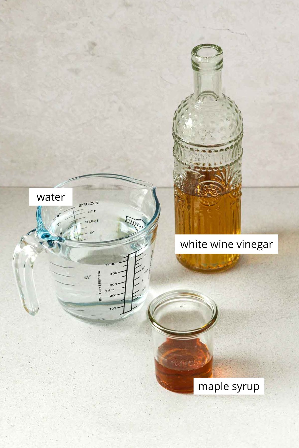 water in glass measuring cup, white wine vinegar and maple syrup in weck jar on grey background with labels.