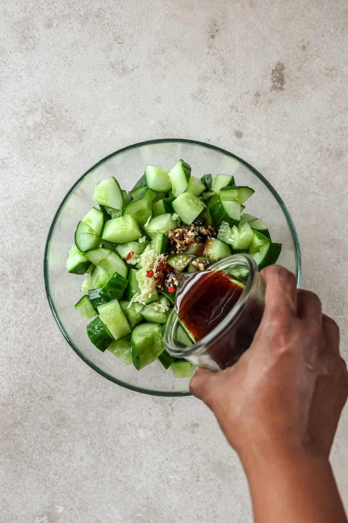 pouring Asian dressing on chopped cucumbers, ginger and chili in glass bowl.