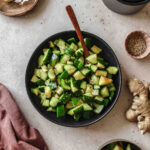 spicy asian smashed cucumber salad on black plate with wooden spoon and props.