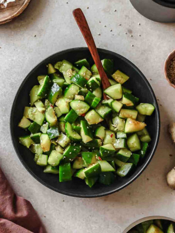 spicy asian smashed cucumber salad on black plate with wooden spoon and props.