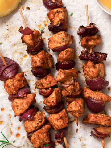 chicken kabobs with red onion on parchment paper with rosemary garnish.