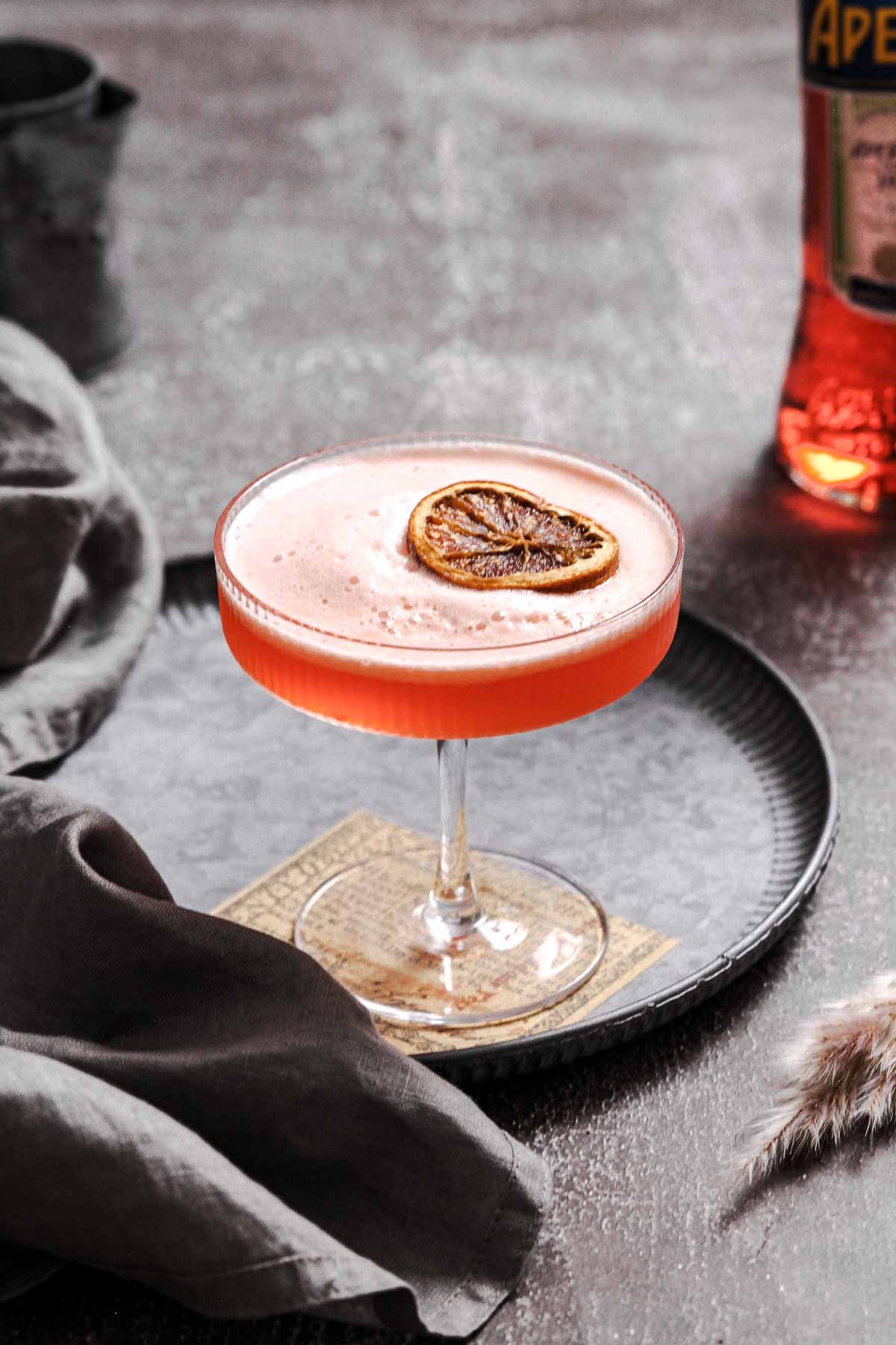 aperol sour aperitif with frothy finish and dried orange slice garnish on vintage plate.