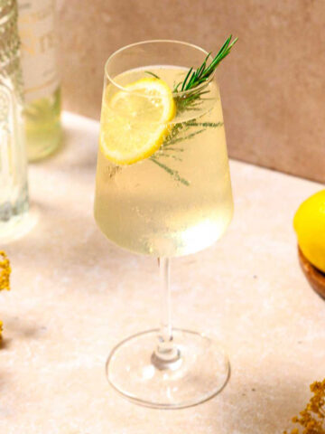 limoncello spritz cocktail with lemon and rosemary garnish on beige marble background.