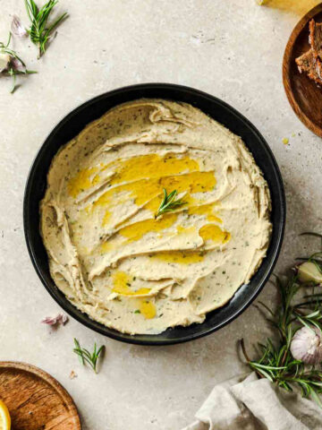 white bean dip with rosemary in black bowl on beige background.