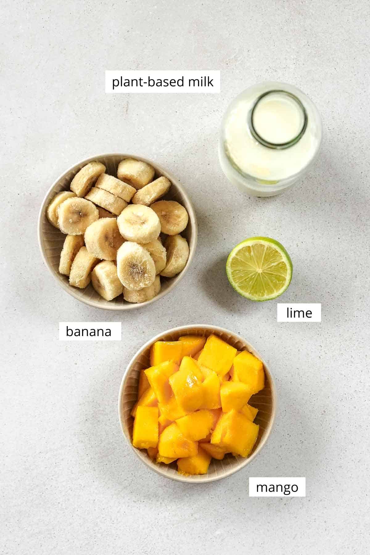 frozen sliced banana, chopped mango, half a lime and plant-based milk on grey background.